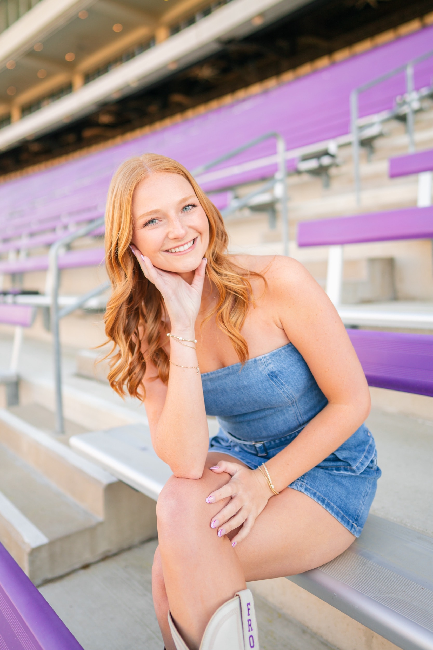 Smiling senior portrait on purple stadium bleachers, wearing a denim outfit in golden light.
