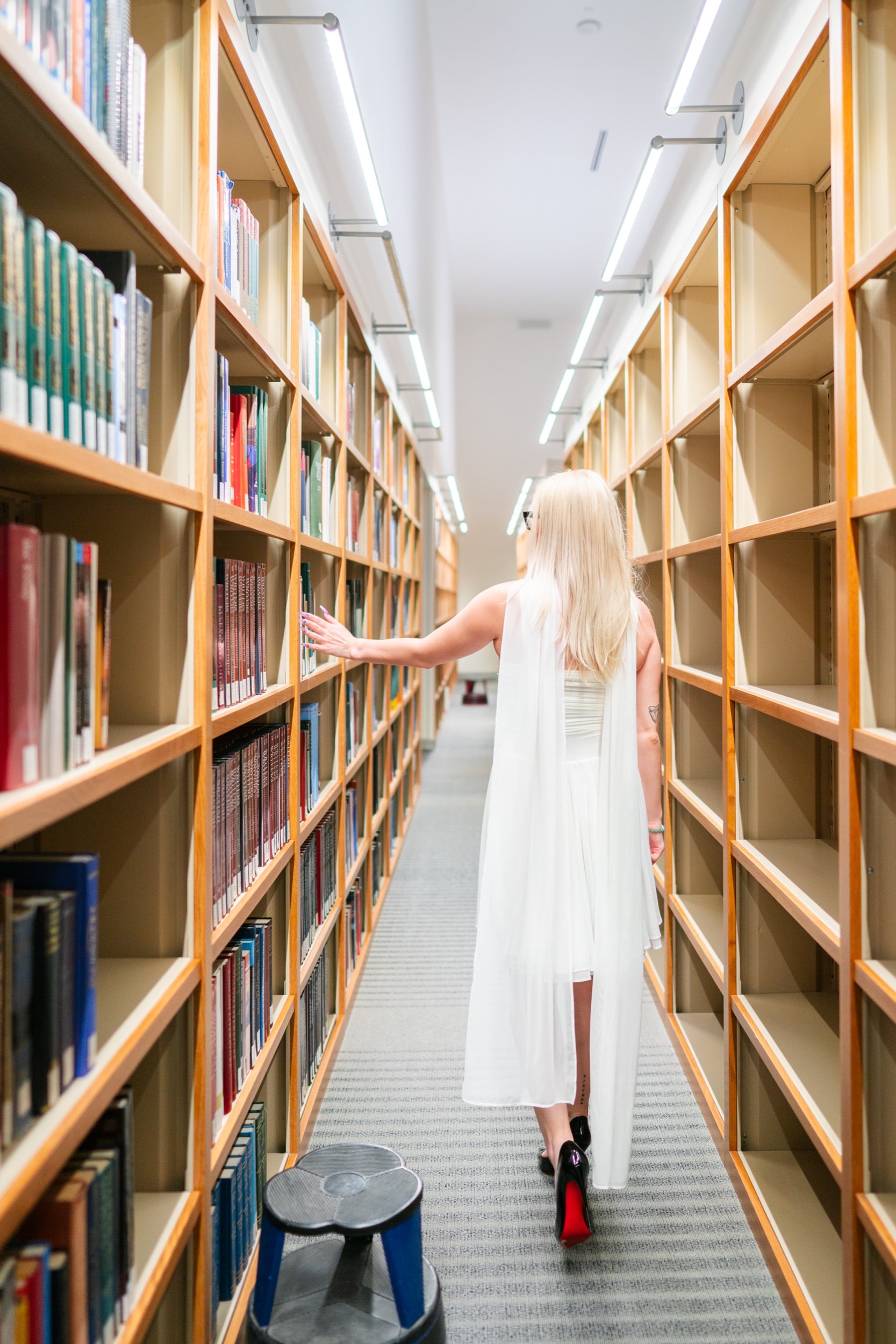 Senior in a flowing white dress walking through library shelves, trailing her hand along books.