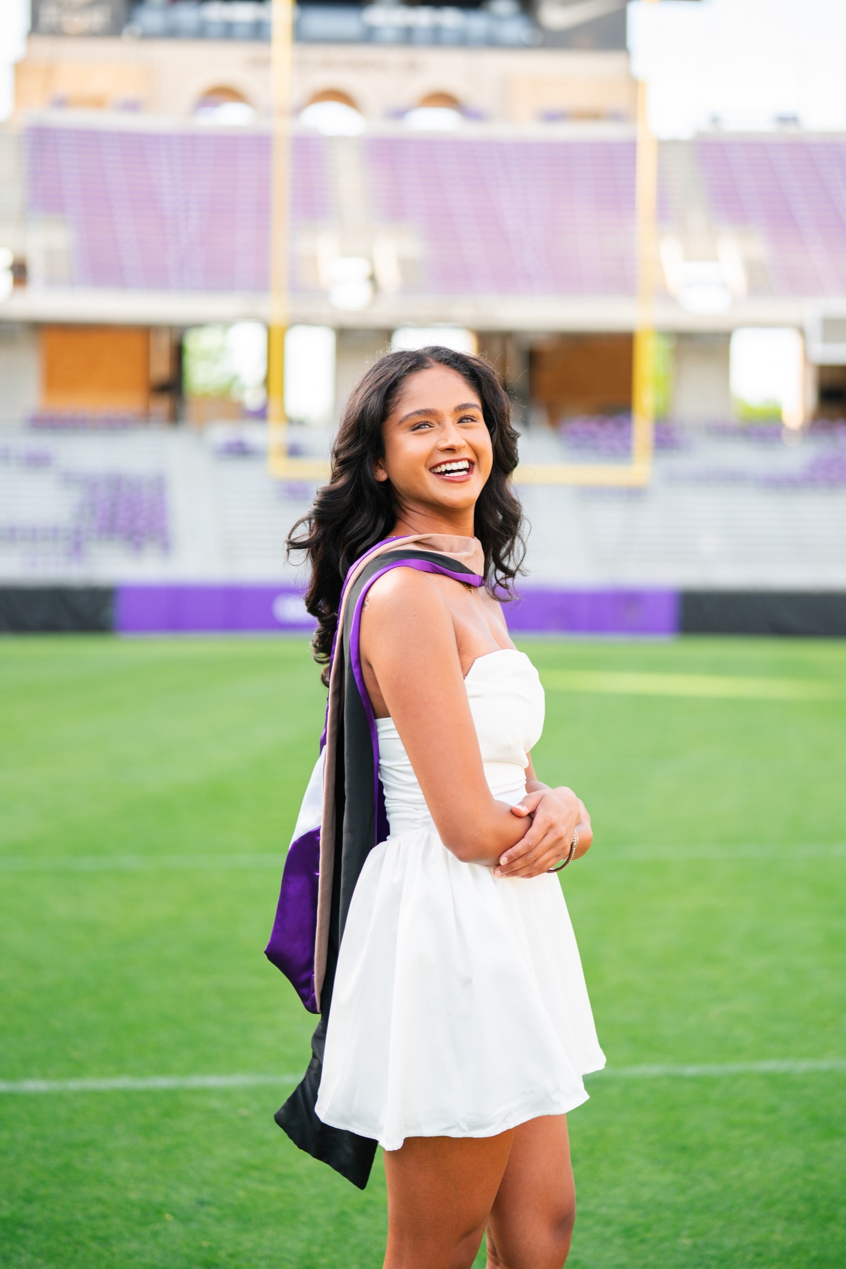 Laughing graduate in a white dress on a football field, graduation stole over her shoulder.