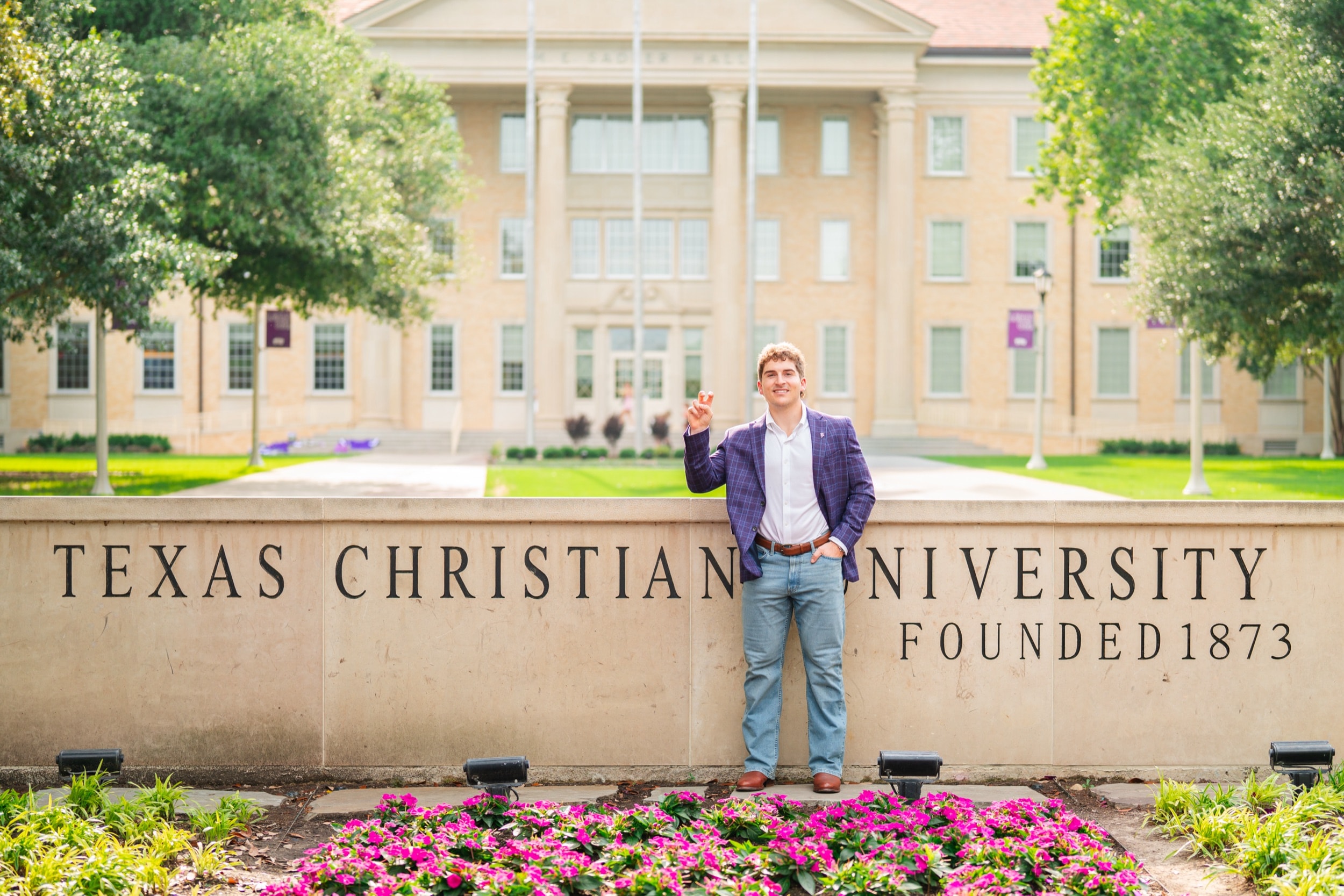 Senior in a blazer smiling in front of a university entrance sign surrounded by flowers.