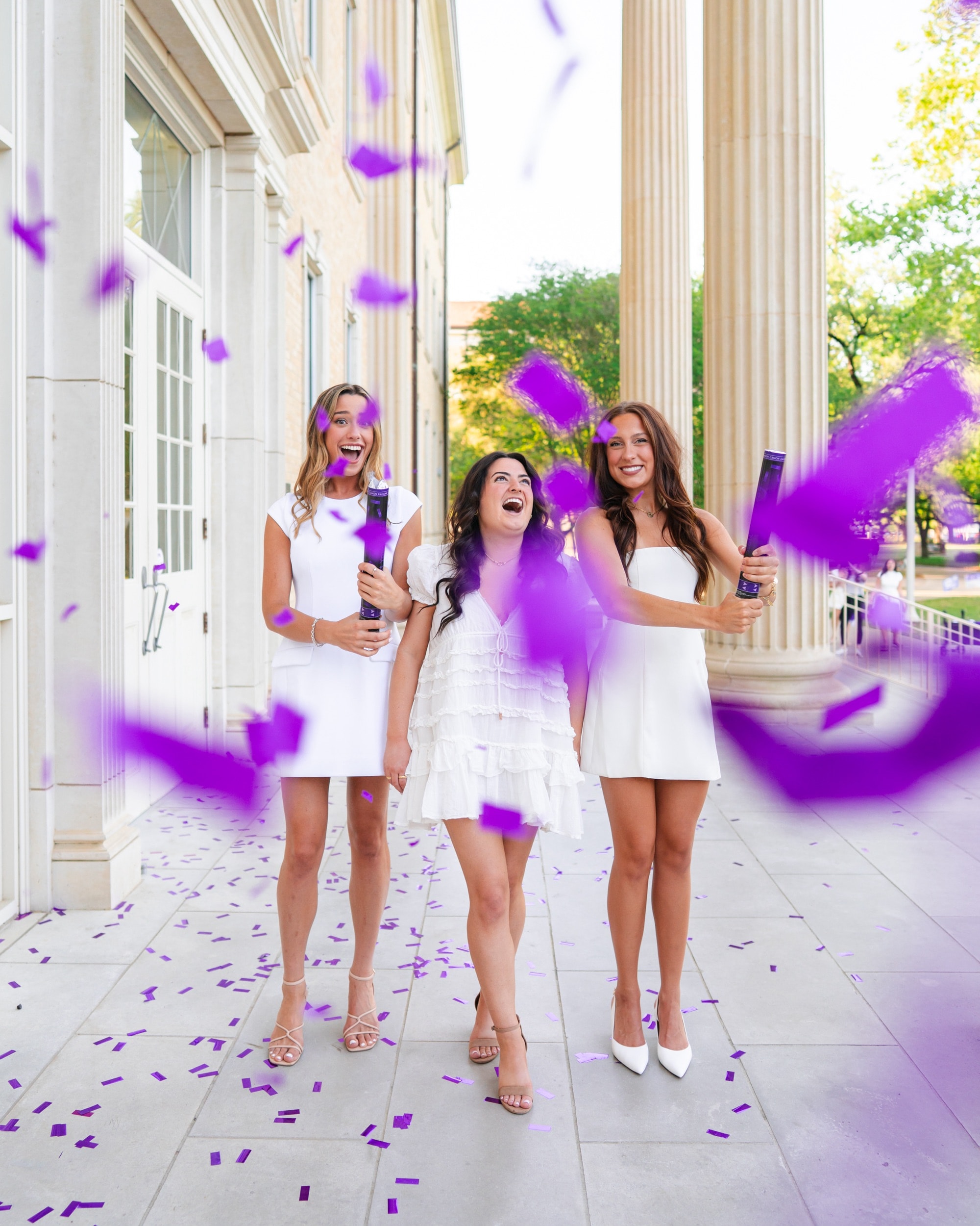 Three friends in white dresses laughing as purple confetti bursts around them outdoors.