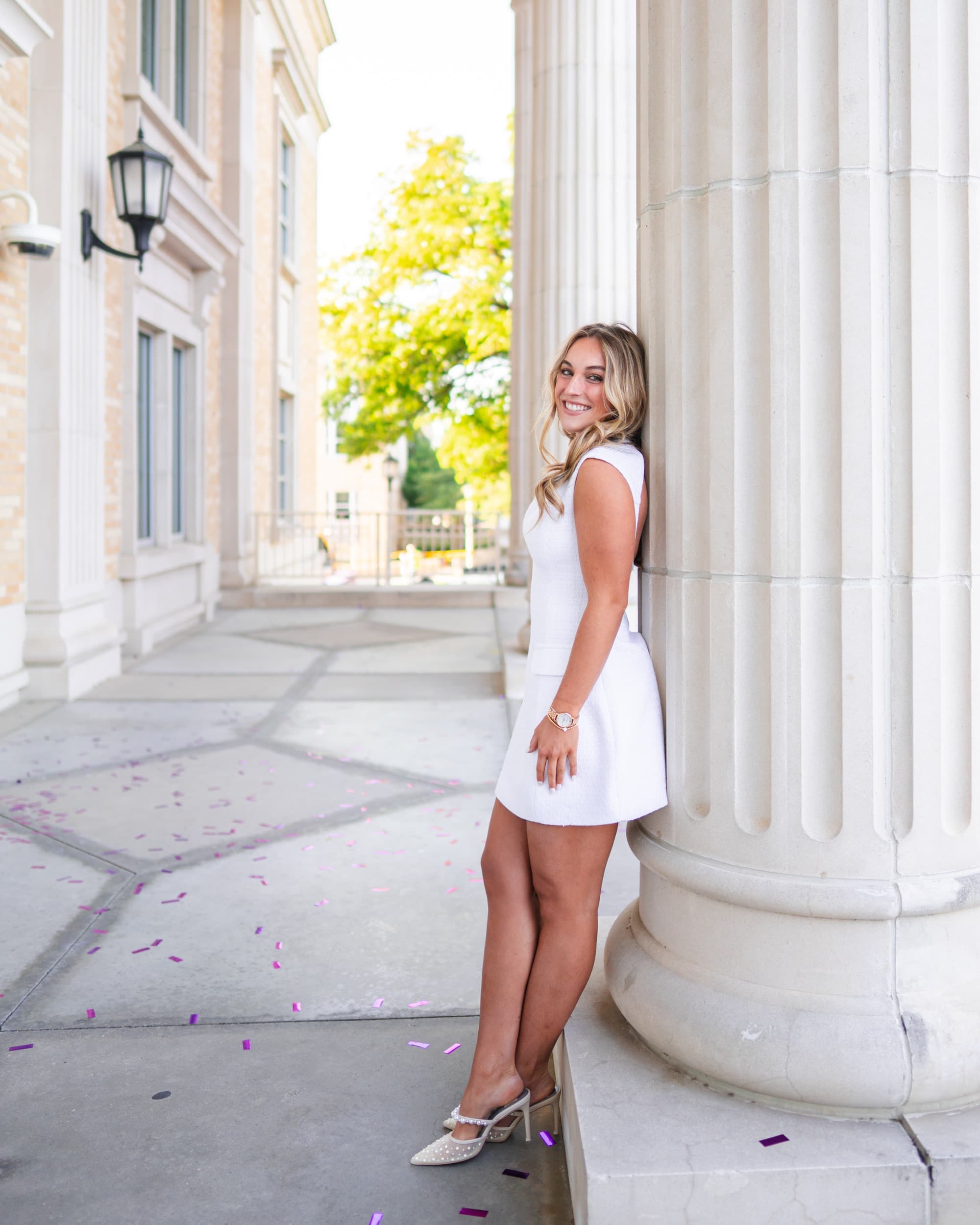 Senior leaning against a stone column in a white dress, glancing back with a smile.