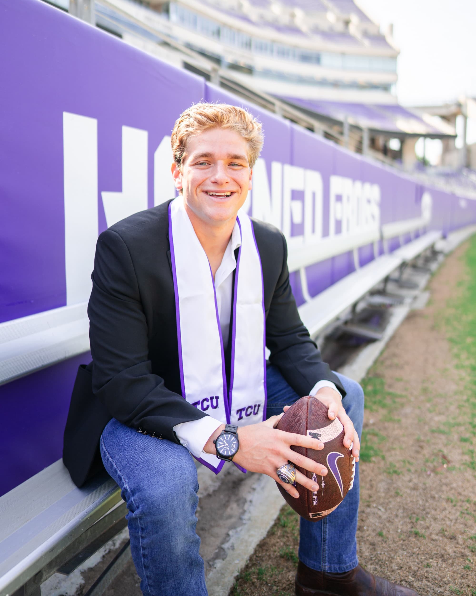 Senior in a blazer holding a football, seated on the stadium wall smiling widely.