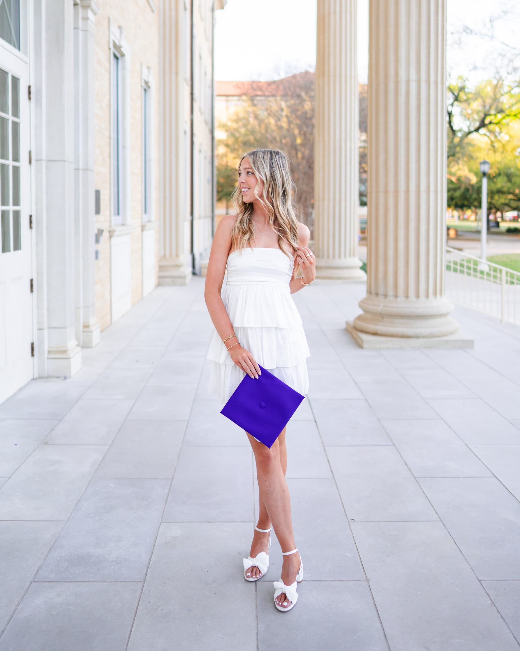Senior in a flowy white dress standing under a colonnade, holding her graduation cap.