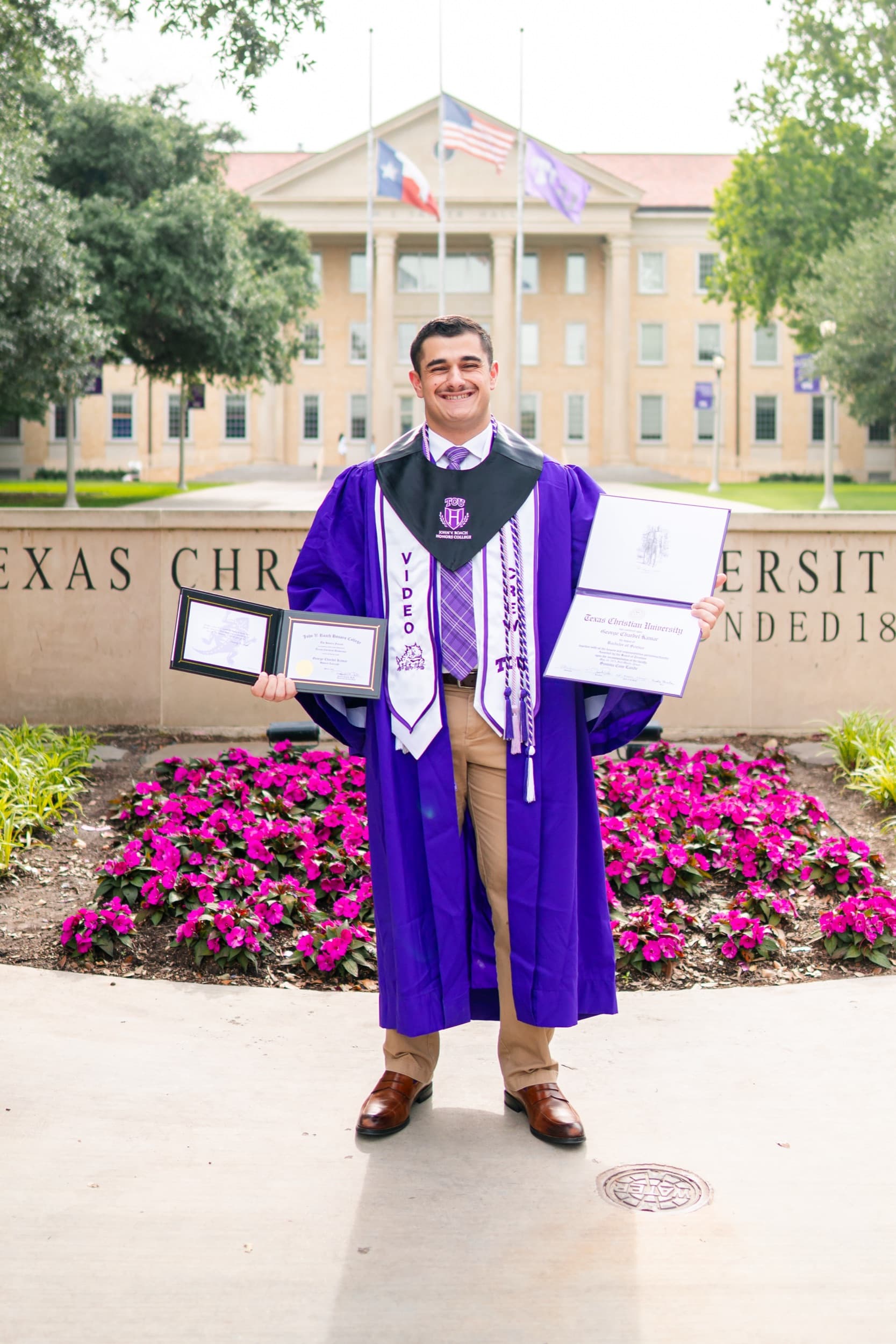 Graduate in a purple gown proudly displaying his diploma in front of a university sign.