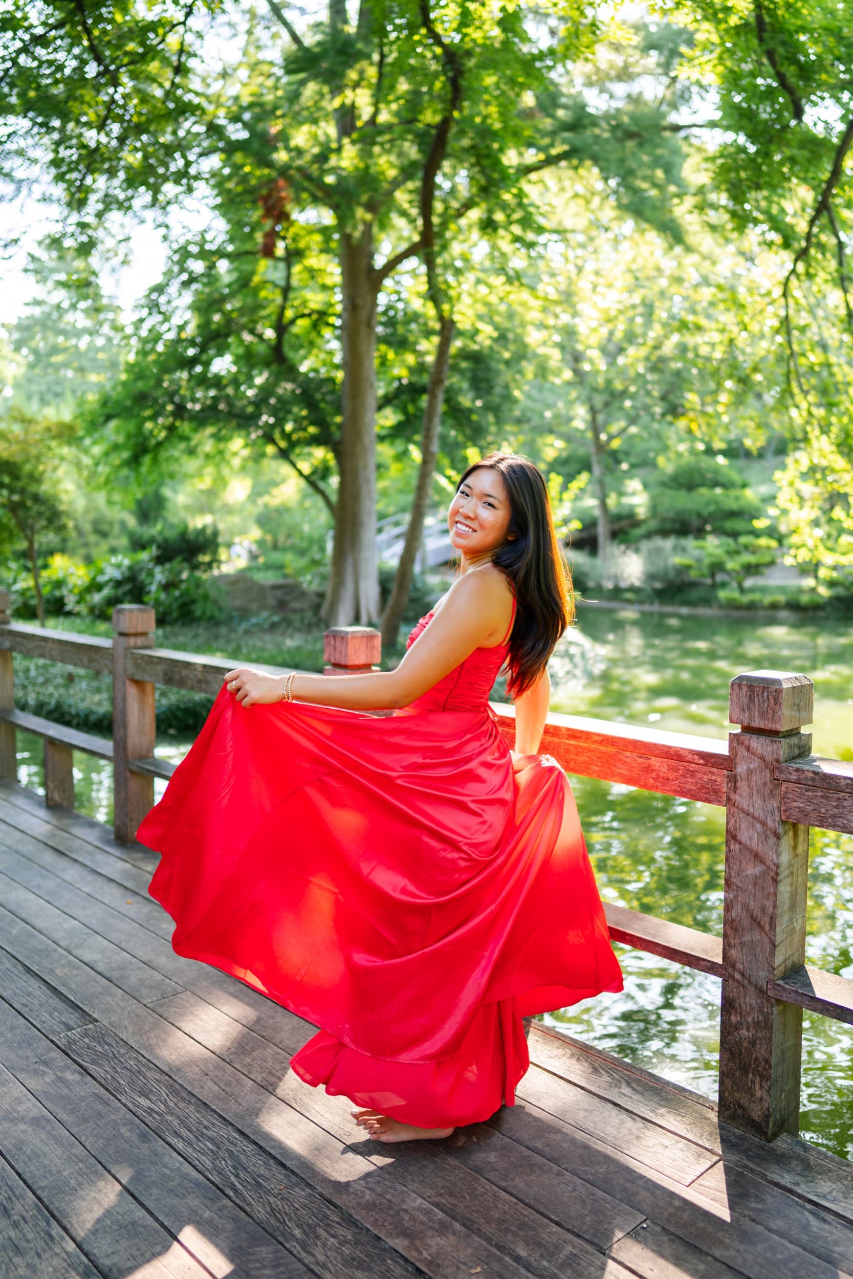Senior twirling in a flowing red gown on a wooden bridge over a garden pond.