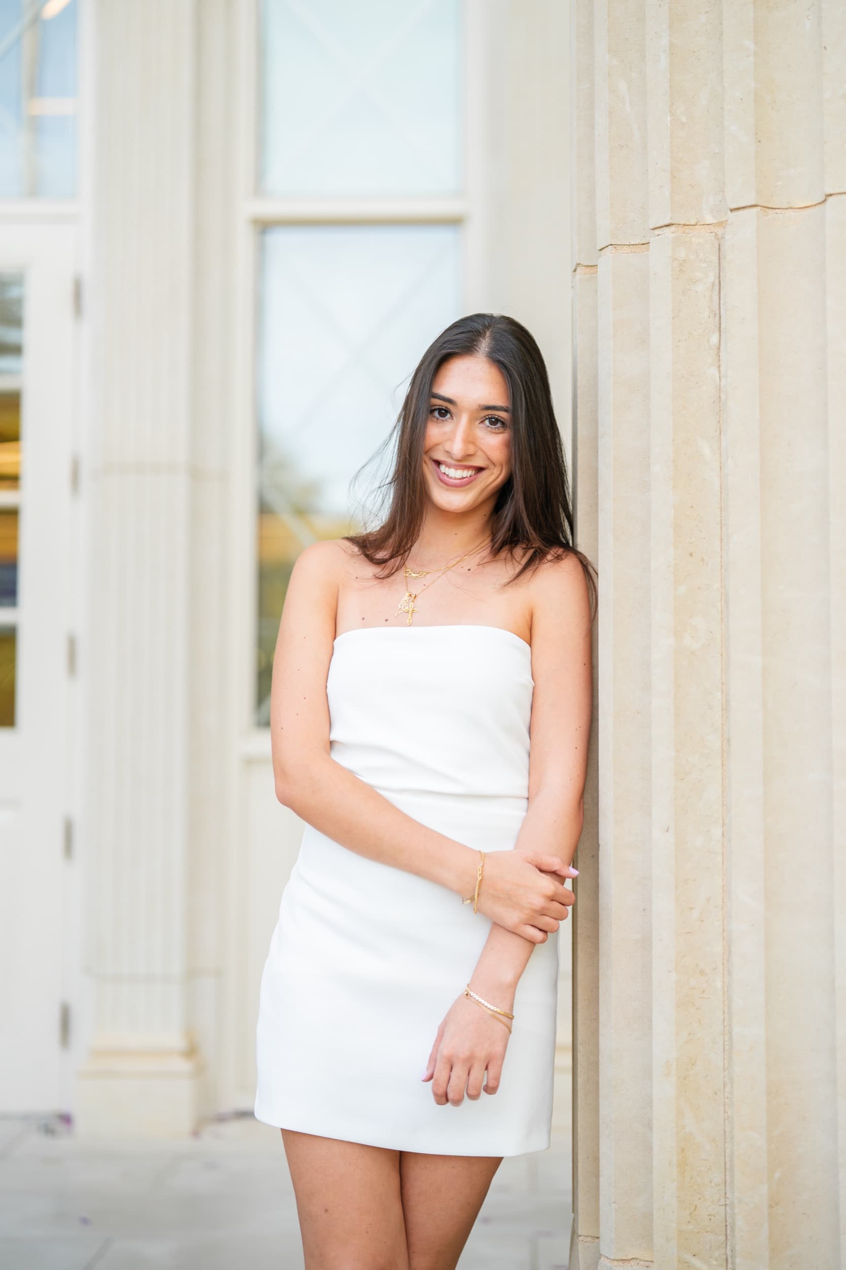 Senior in a white strapless dress leaning against a sunlit stone column, smiling warmly.