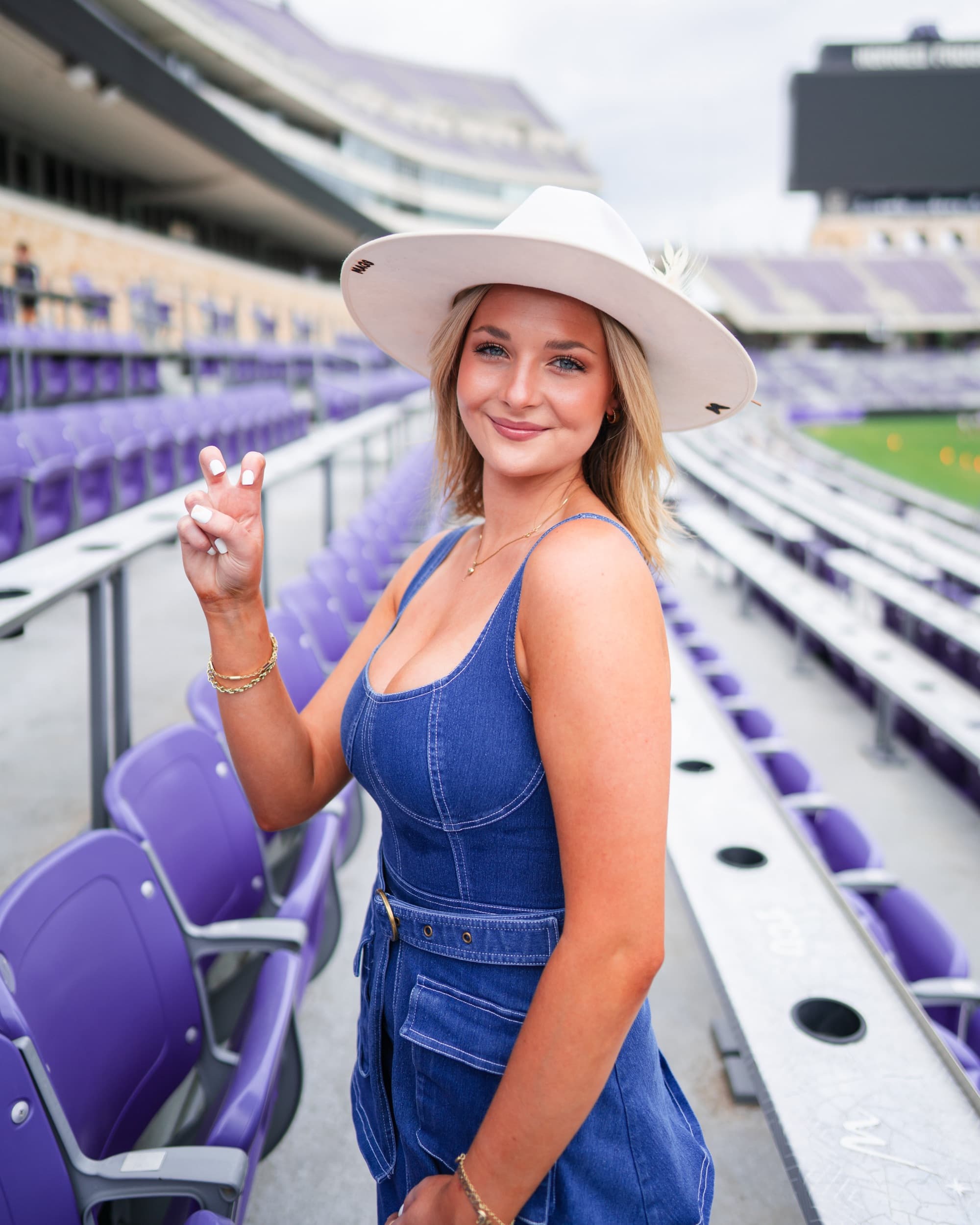 Senior in a wide-brim hat and denim overalls smiling in an empty stadium.