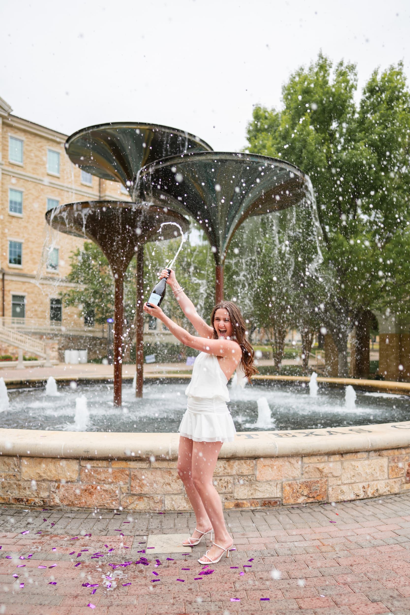 Senior in a white dress popping champagne in celebration in front of a fountain.