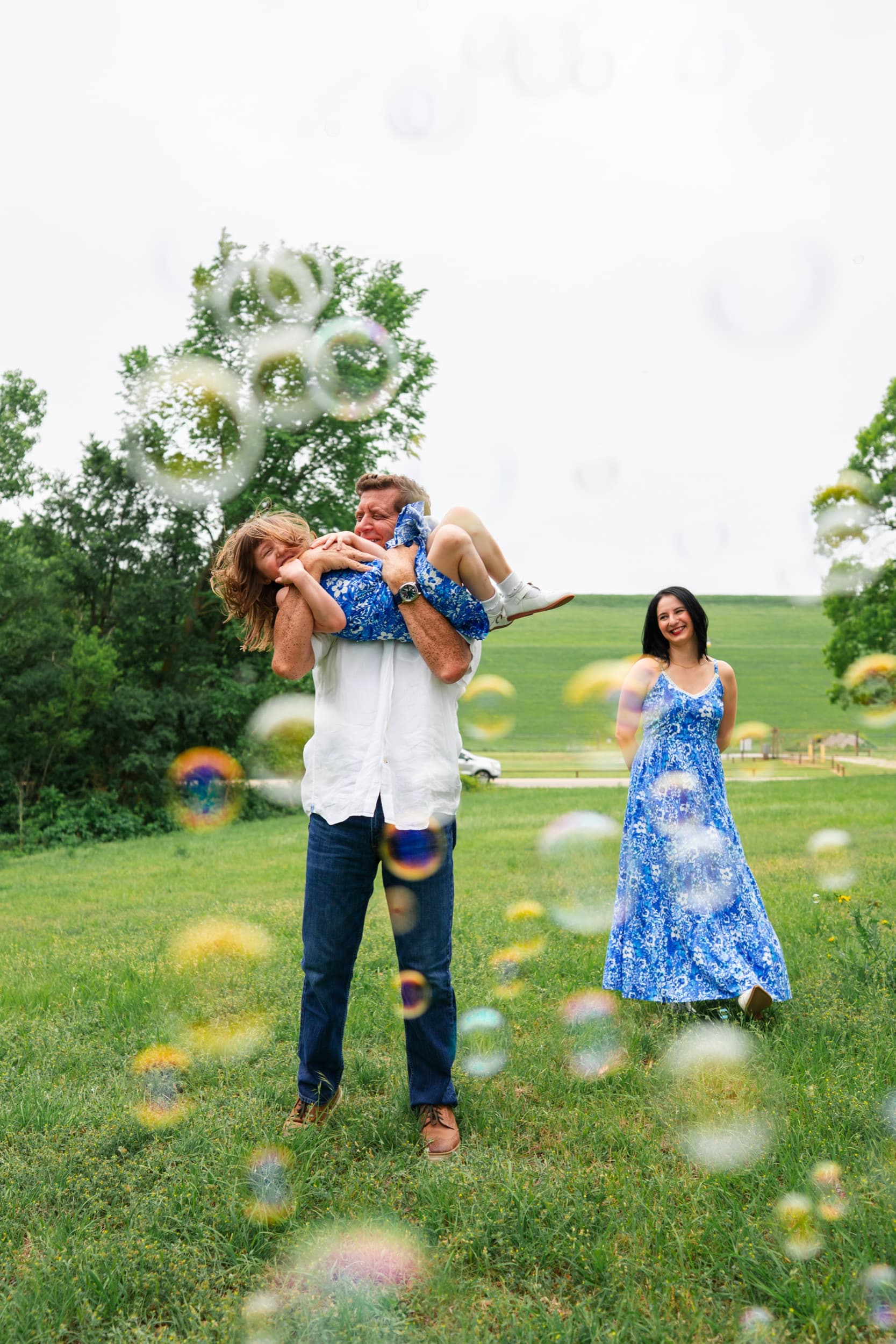 Father playfully lifting a laughing child while mom watches, bubbles floating in a grassy field.