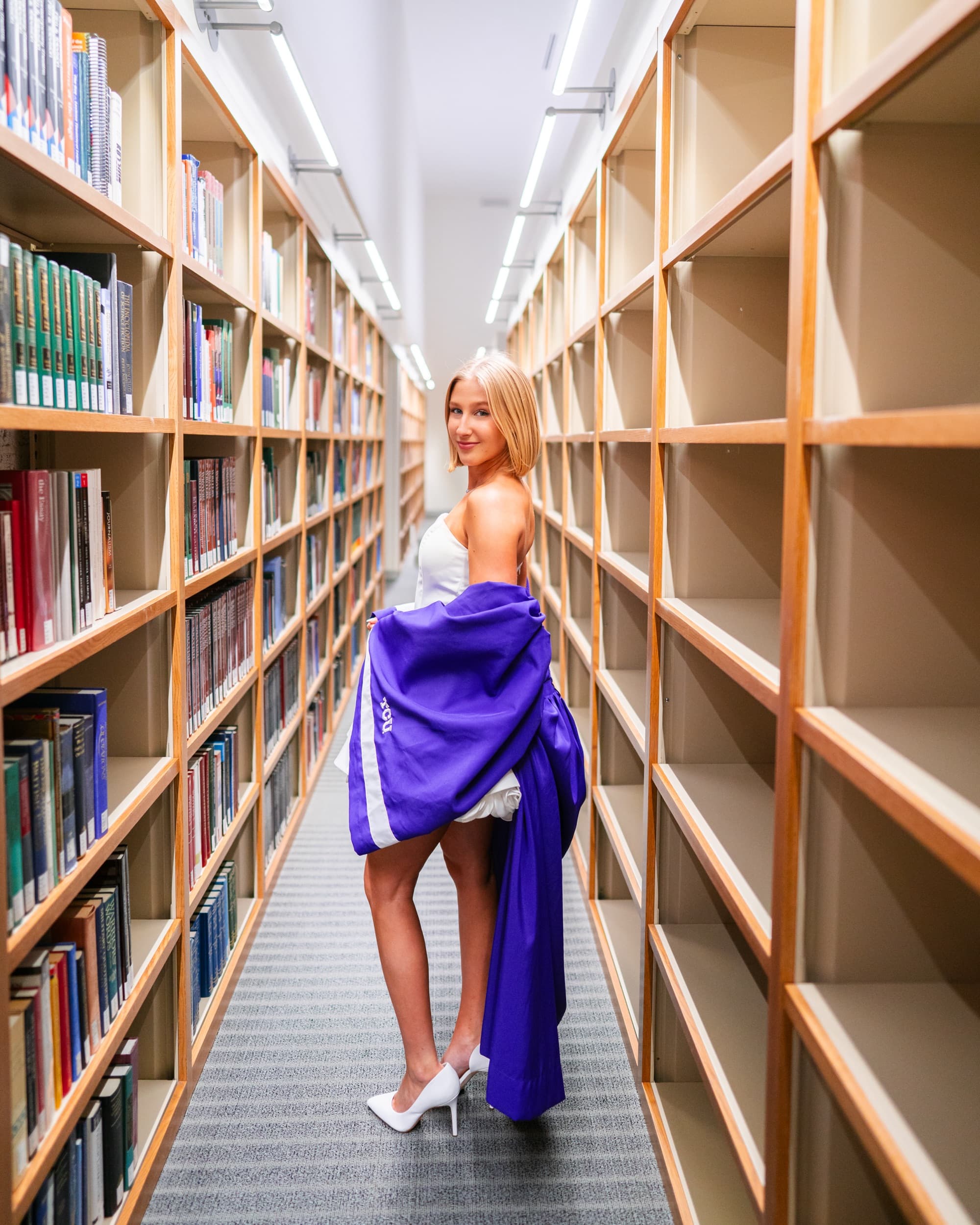 Senior in a white dress holding a purple graduation gown in a library aisle.